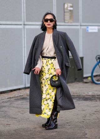 Pornwika Spiecker wears sunglasses, earrings, a gray lg coat, a white wool cardigan, a yellow floral print long skirt , a Miu Miu leather bag, pointed leather boots, outside TG Botanical, during the Copenhagen Fashion Week AW24 on February 01, 2024 in Copenhagen, Denmark.