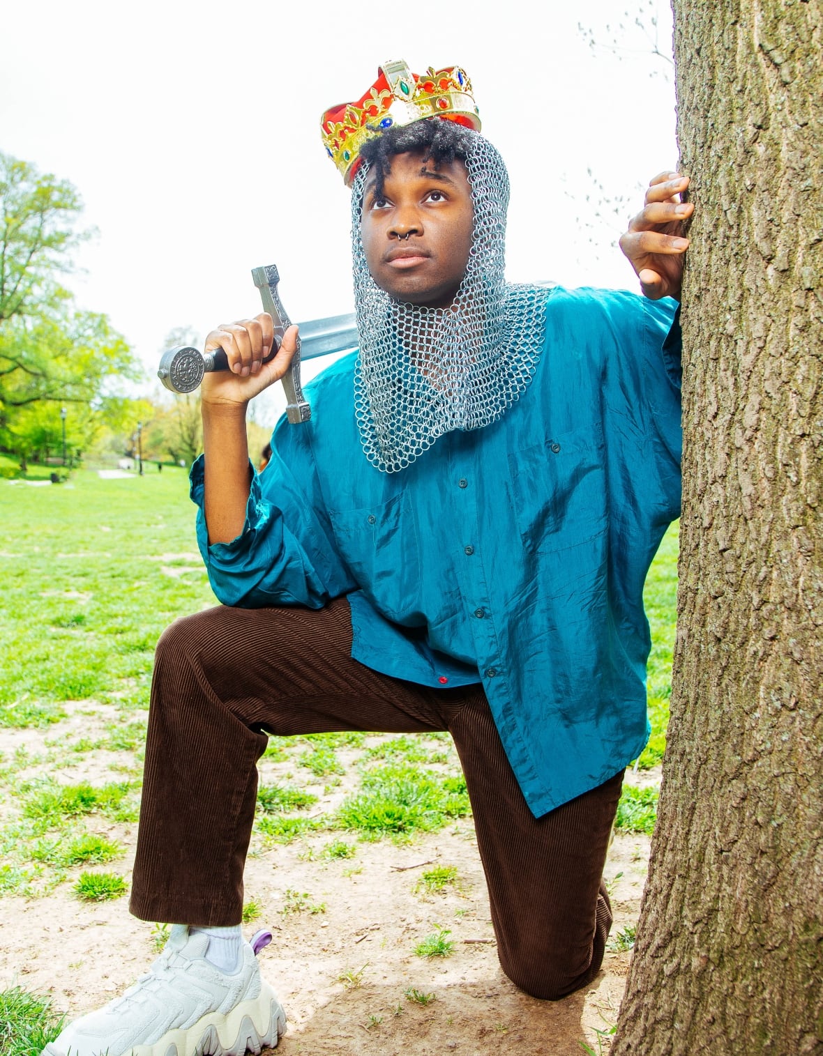A person wearing a stylized crown and chainmail coif kneels with a knee down, one hand bracing against a tree as the other balances an imitation sword on their shoulder.