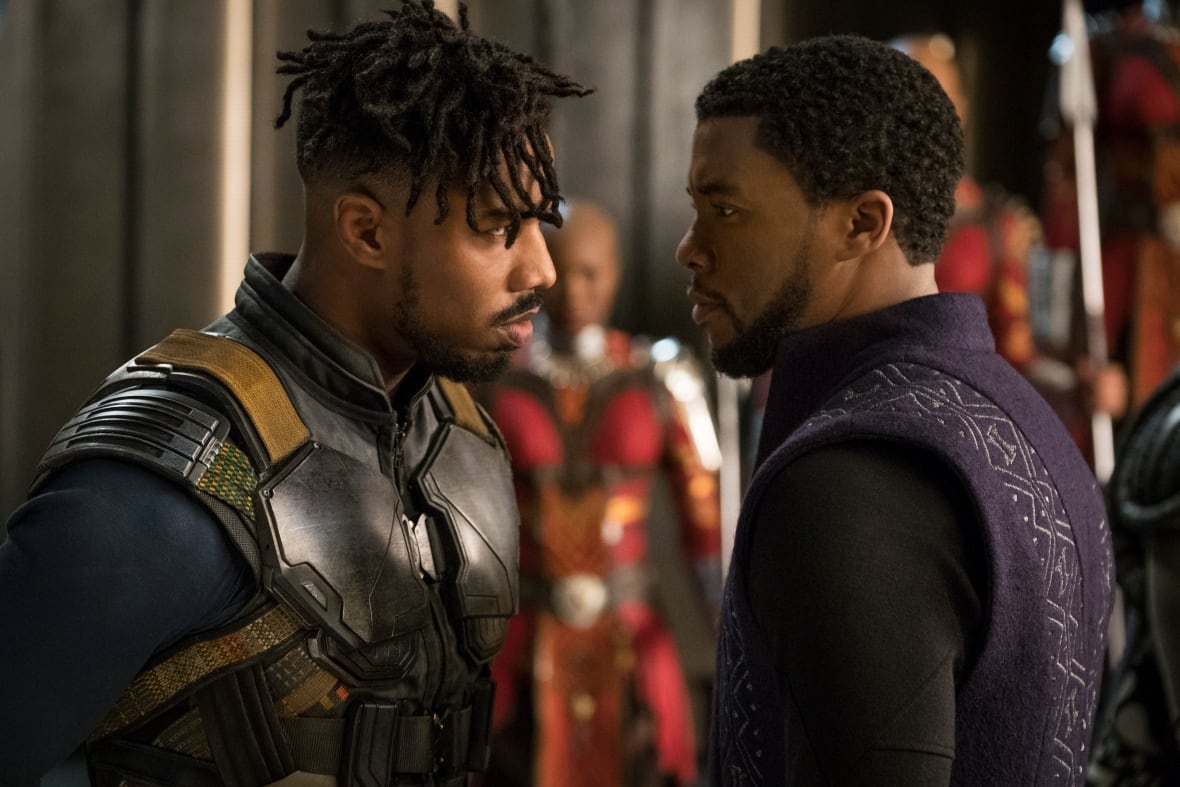 A close-up image of two men standing close to each other and staring intently in a face-off. Women in vibrant armour are seen out of focus in the background.