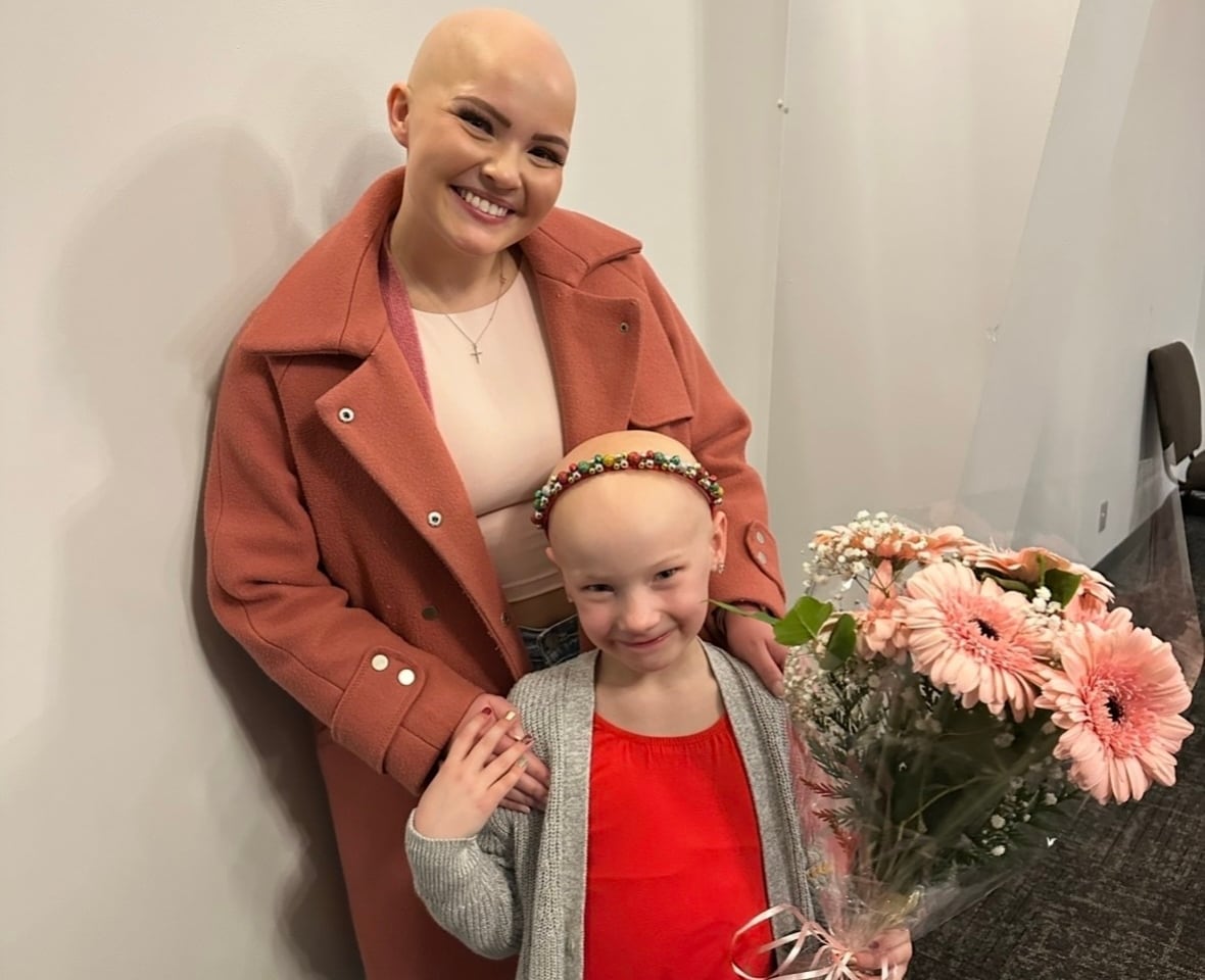 A woman and a girl, both bald, pose for a photo. The girl holds a bouquet of pink flowers.