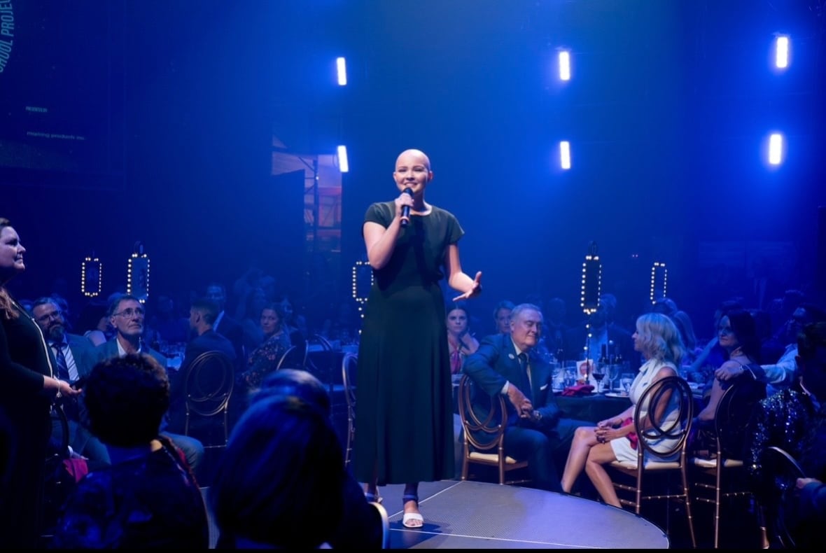 A young woman in a dark dress stands on a circular platform holding a mic, surrounded by people sitting at tables.