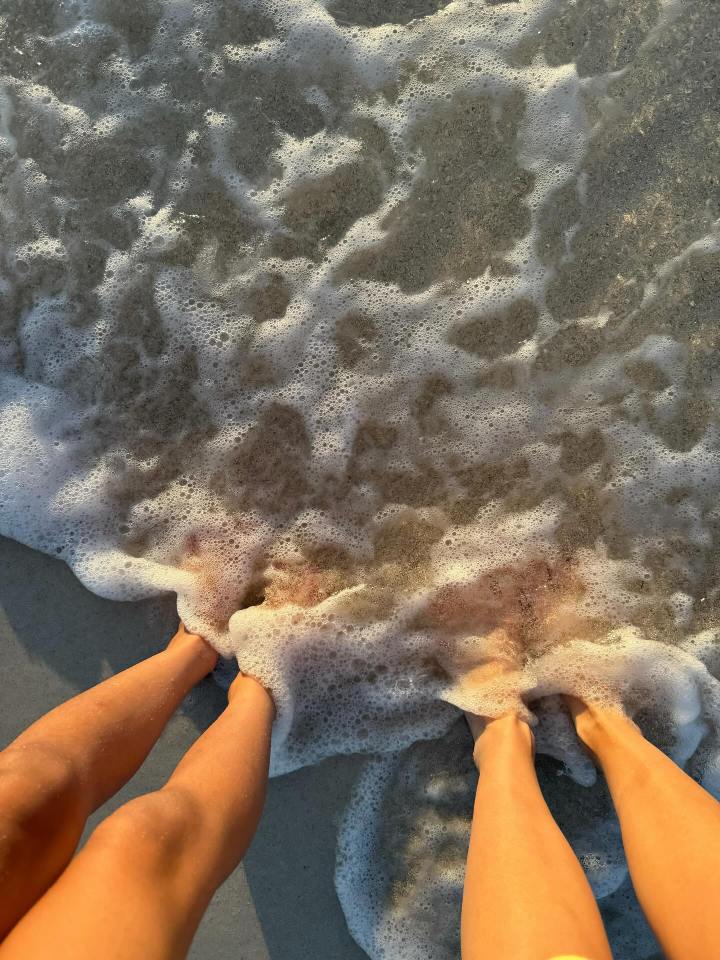 Two pairs of legs in ocean foam on a sandy beach.